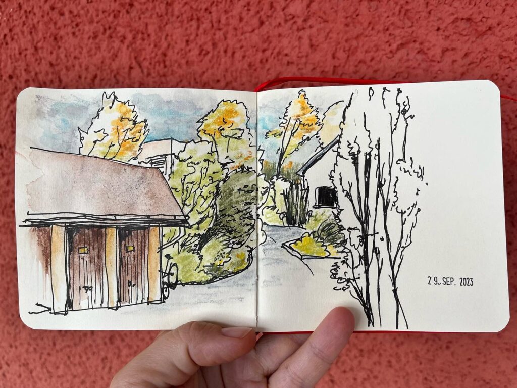 Aufgeschlagenes Skizzenbuch mit Tusche- und Aquarellzeichnung eines Dorfwegs mit Häusern und gelben Bäumen im Herbst.