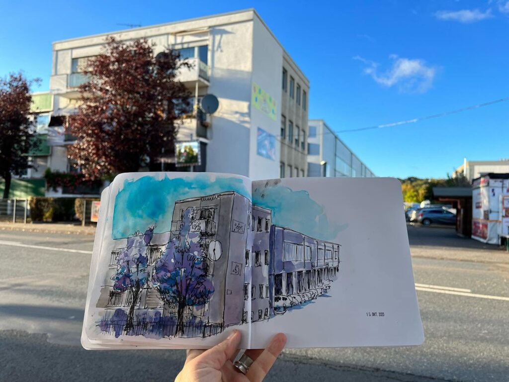 Ein aufgeschlagenes Skizzenbuch mit Stadtzeichnung wird vor der gezeichneten Straße gehalten.